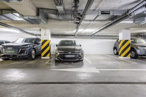 A parking garage with three cars parked in designated spaces, concrete pillars with yellow and black stripes, and overhead pipes. - Apartment near River Walks (Warszawa 2)