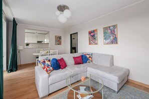 Un salon moderne avec un canapé d’angle gris, des coussins colorés, une table basse en verre et une cuisine en arrière-plan.
