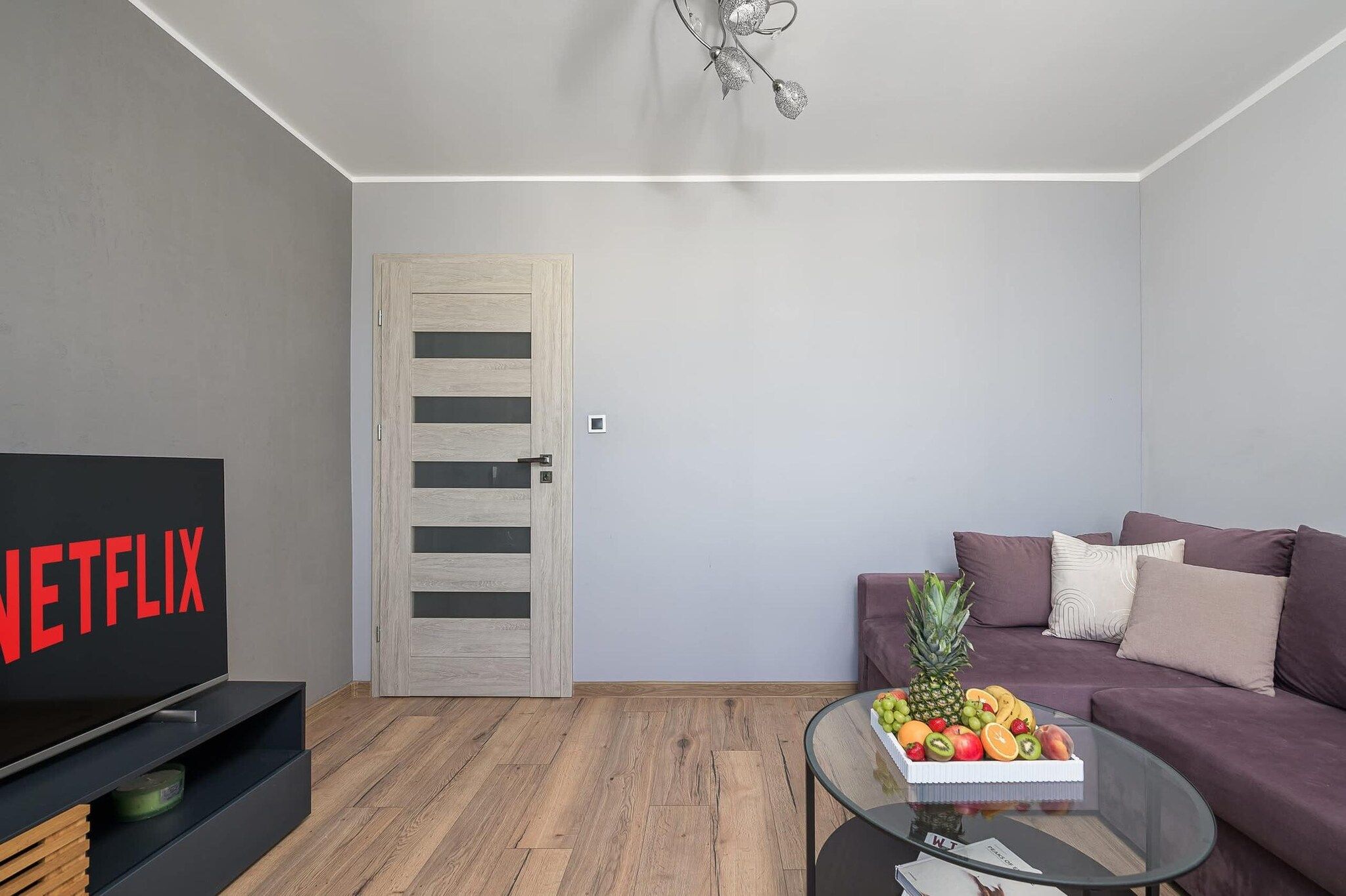 Una sala de estar moderna con un televisor de pantalla plana, una puerta de madera, una mesa de centro de cristal con fruta y un sofá morado.