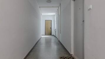 Un pasillo largo y estrecho con paredes blancas y suelo de hormigón. Hay una puerta al final del pasillo, y las paredes tienen una serie de ventanas pequeñas y regularmente espaciadas.