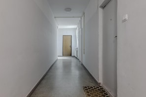 Un pasillo largo y estrecho con paredes blancas y suelo de hormigón. Hay una puerta al final del pasillo, y las paredes tienen una serie de ventanas pequeñas y regularmente espaciadas.