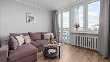 Una sala de estar moderna con un sofá modular, una mesa de centro de vidrio con frutas y vistas de la ciudad a través de amplias ventanas.