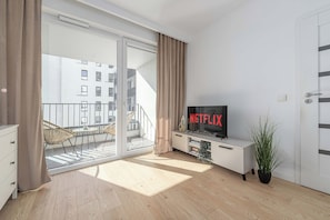 A modern living room with a large TV displaying 'NETFLIX', a sliding glass door leading to a balcony, and a potted plant. - Retreat by Park Skaryszewski (Warszawa)