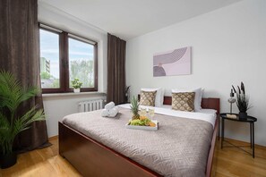 Ein Hotelzimmer mit einem Bett, einem Vorhangfenster, einem Heizkörper, einem kleinen Tisch mit einer Pflanze und einem an der Wand hängenden Kunstwerk.