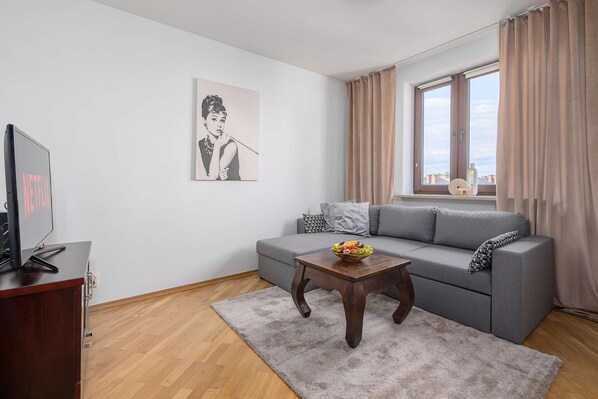 Ein modernes Wohnzimmer mit einem grauen Sofa, einem Couchtisch mit Obst, einem Fernseher auf einem Schrank und einem Fenster mit Vorhängen.