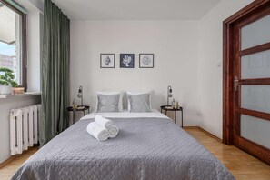 A neatly arranged bedroom with a bed, two bedside tables, a radiator, and a window with curtains. - Urban Oasis with Balcony near Zoo (Warszawa)