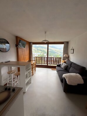 Un salon avec un canapé, un coin repas avec une table et des chaises, un coin cuisine avec une cuisinière, et une grande fenêtre donnant sur des collines.