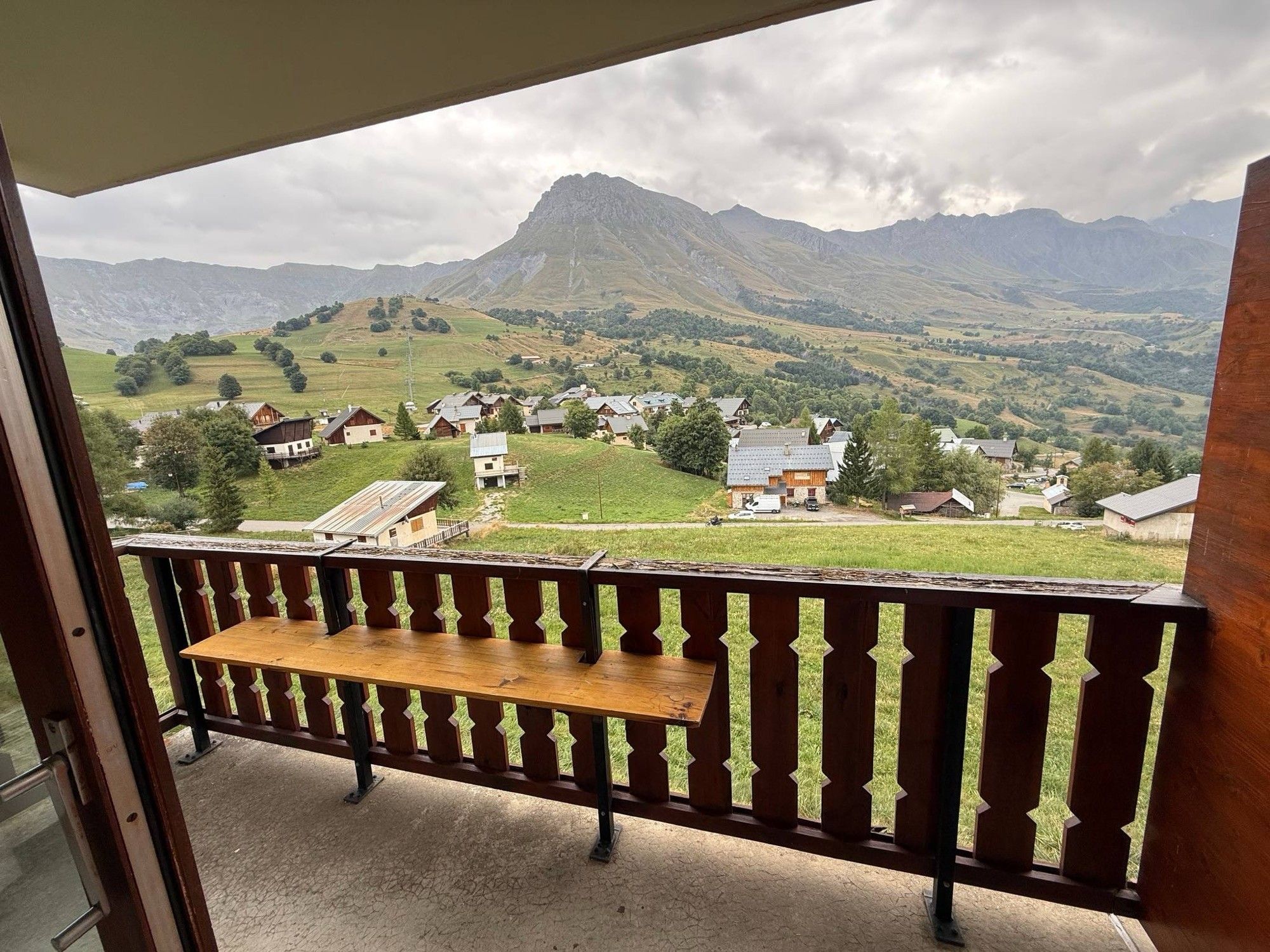A balcony with a wooden bench, overlooking a village with houses and a mountain in the background.