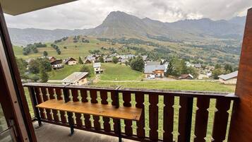 A balcony with a wooden bench, overlooking a village with houses and a mountain in the background.