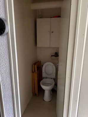 A small, narrow bathroom with a toilet, a wooden shelf, and a white cabinet.