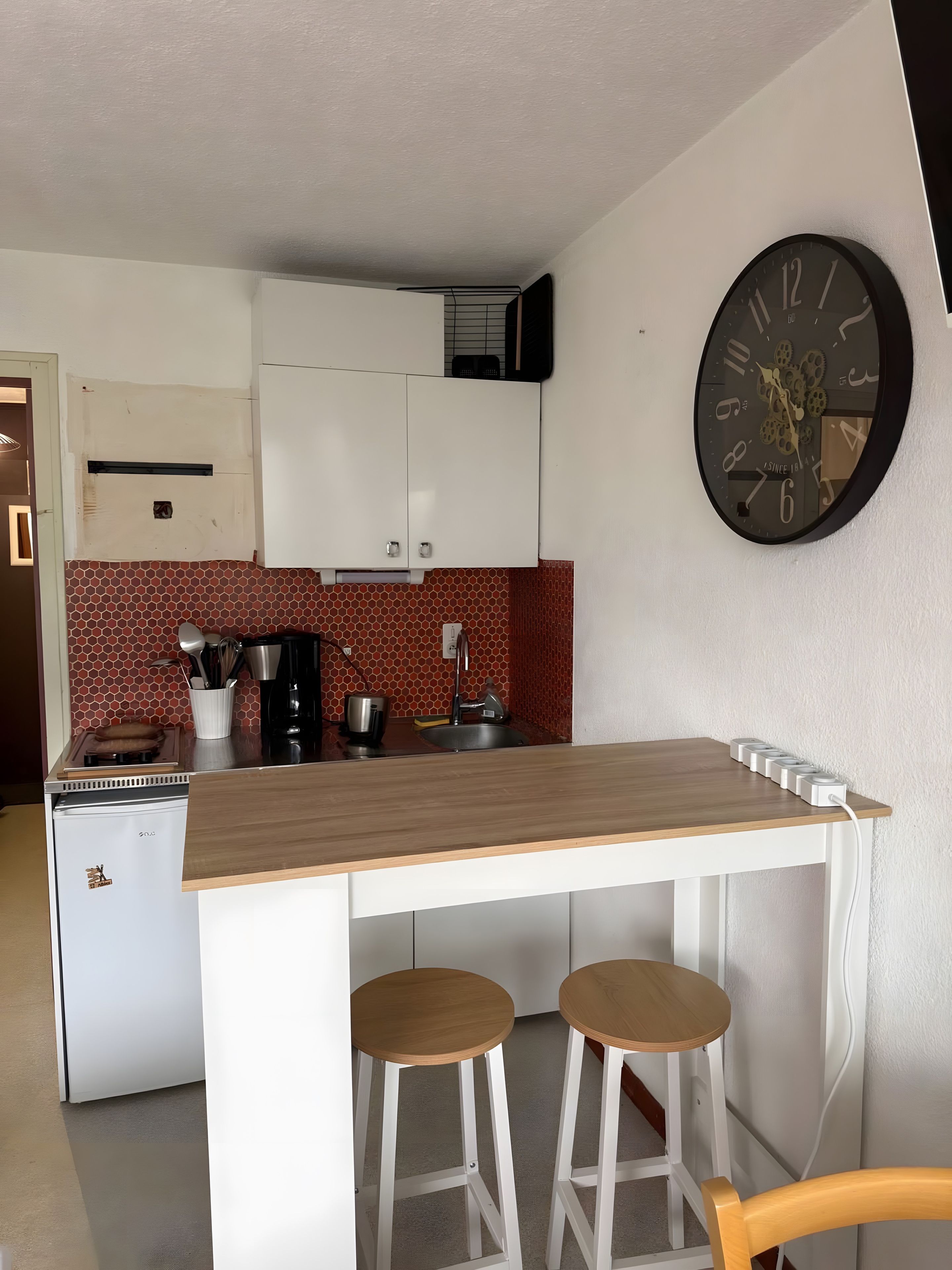 A compact kitchen with a wooden countertop, two stools, a wall clock, and a small refrigerator.