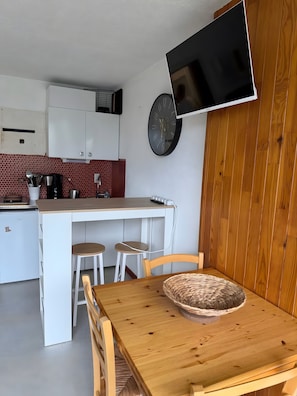 Une cuisine compacte avec des meubles blancs, une table à manger en bois et un téléviseur fixé au mur.