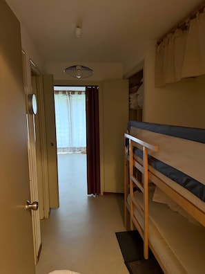 Une pièce avec un lit superposé, une porte dotée d’une poignée ronde, un luminaire au plafond et une vue sur un espace extérieur, visible à travers un rideau.