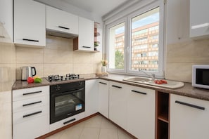 A modern kitchen with white cabinets, a gas stove, an oven, a sink, and a window with a view of buildings. - Riverside Flat with Balcony (Warszawa)