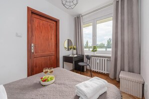 Habitación de hotel con puerta de madera, escritorio con silla, cama con colcha estampada, mesita con frutero y vasos, y ventana con vistas al exterior.