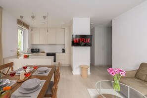 Une cuisine moderne avec un coin repas, un téléviseur à écran plat fixé au mur et un salon comprenant un canapé et des fleurs.
