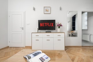 Ein modernes Wohnzimmer mit einem Flachbildfernseher, der 'NETFLIX' anzeigt, einem weißen Schrank mit Schubladen, einer Zeitschrift auf dem Boden und einer Tür links.