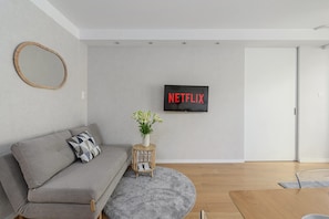 A modern living room with a grey sofa, a small side table with a plant, a round rug, and a wall-mounted TV displaying 'NETFLIX'. - Saska Apartment near Old Town (Warszawa)
