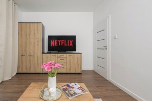 Een moderne woonkamer met een houten kast, een televisie waarop 'NETFLIX' wordt weergegeven, een vaas met roze bloemen en een tijdschrift op een houten tafel.