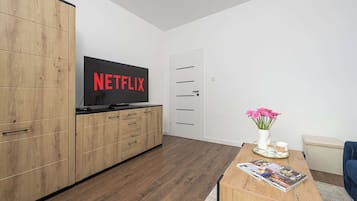 Un salon moderne avec un téléviseur à écran plat, des meubles en bois, une table basse avec des magazines, un vase rempli de fleurs et un canapé bleu.