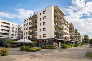 Un immeuble résidentiel à plusieurs étages, doté de balcons, d’une cour pavée et d’un espace extérieur aménagé pour les cafés.