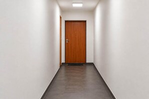 Un couloir se terminant par une porte en bois, avec des murs blancs et un sol carrelé.