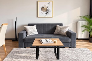 A modern living room with a grey sofa, two decorative pillows, a wooden coffee table with magazines and a candle, a framed artwork above the sofa, and a potted plant to the right. - Apartment near Old Town & Zoo (Warszawa)