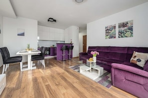 A modern living room with a purple sofa, a dining table with chairs, a kitchen area, and artwork on the walls. - Cozy Apartment with Skyline View (Warszawa)