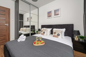 A hotel room with a large bed, a mirror, two framed pictures, a bedside table with a lamp, and a tray of fruit and champagne glasses. - Cozy Apartment with Skyline View (Warszawa)