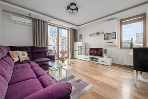 A modern living room with a purple sofa, a glass coffee table, a flat-screen TV, and a city view through large windows. - Cozy Apartment with Skyline View (Warszawa)