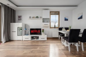 A modern living room with a flat-screen TV displaying 'NETFLIX,' a dining area with a white table and black chairs, and a window with a view of buildings. - Cozy Apartment with Skyline View (Warszawa)