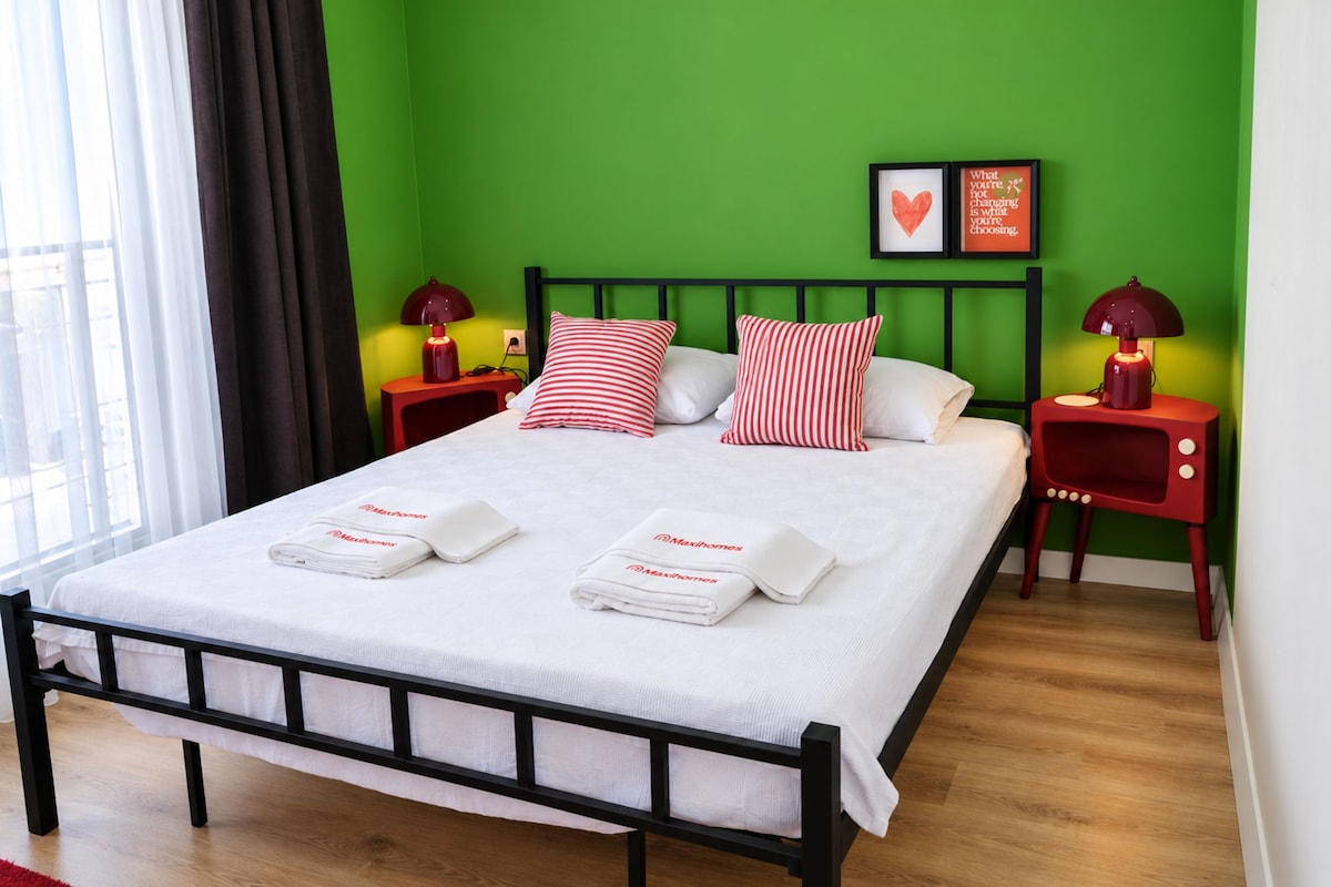 A neatly made bed with white linens and red and white striped pillows, two bedside tables with lamps, and a green wall with a framed picture.