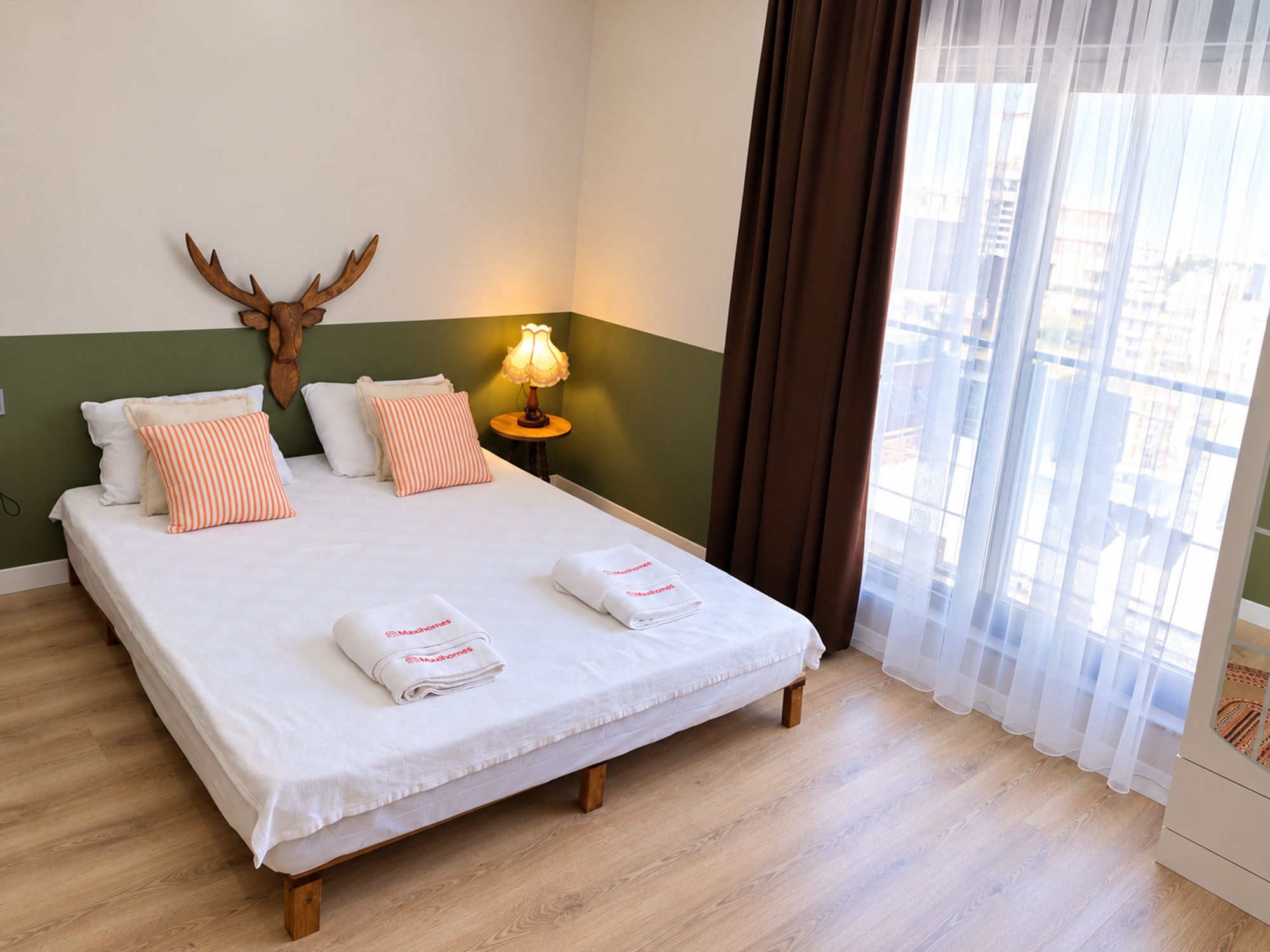 A hotel room with a bed, pillows, a lamp, a deer's head decoration, and a window with curtains.