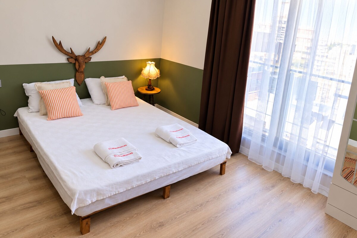 A hotel room with a bed, pillows, a lamp, a deer's head decoration, and a window with curtains.