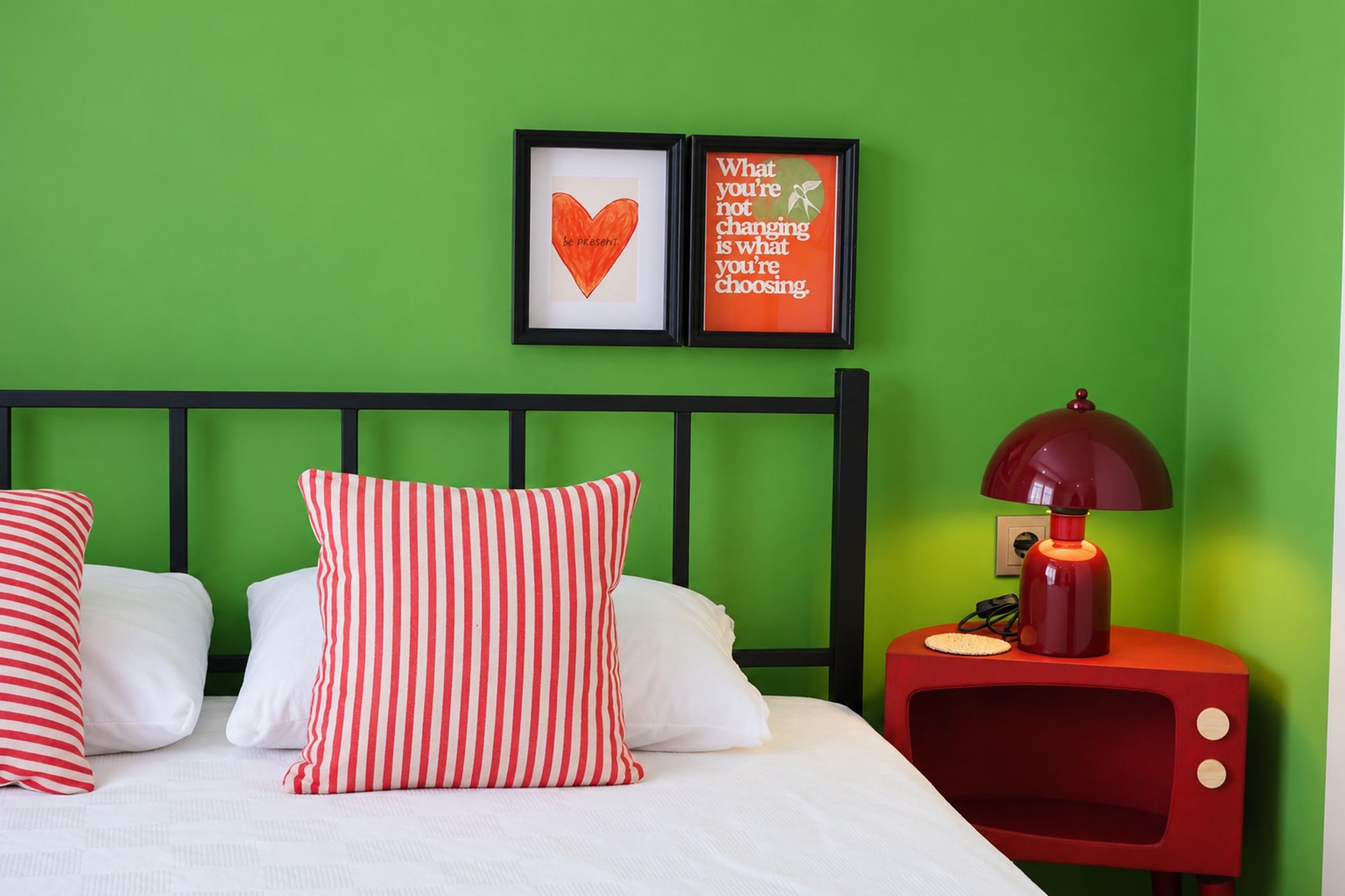 A room with a green wall, a bed with white bedding and striped pillows, a red nightstand with a lamp, and two framed motivational quotes.
