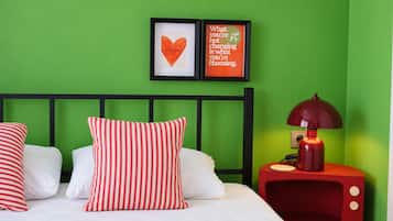 A room with a green wall, a bed with white bedding and striped pillows, a red nightstand with a lamp, and two framed motivational quotes.