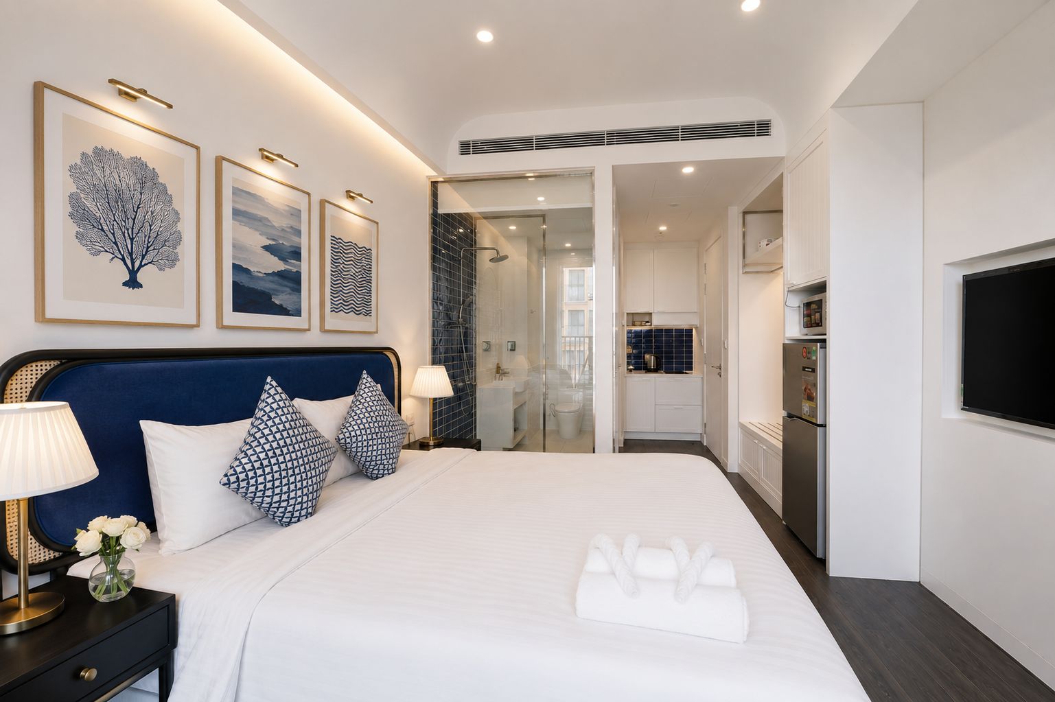 A hotel room with a bed, bedside lamps, a TV, a bathroom with a shower, and a kitchenette.