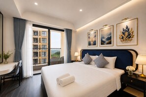 A hotel room with a large bed, a desk, and a view of a building through the window. - Rei Gallery Appartments Hillside (Phu Quoc)