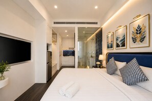 A modern hotel room with a large bed, a flat-screen TV, a walk-in closet, and a bathroom with a glass shower enclosure. - Rei Gallery Appartments Hillside (Phu Quoc)