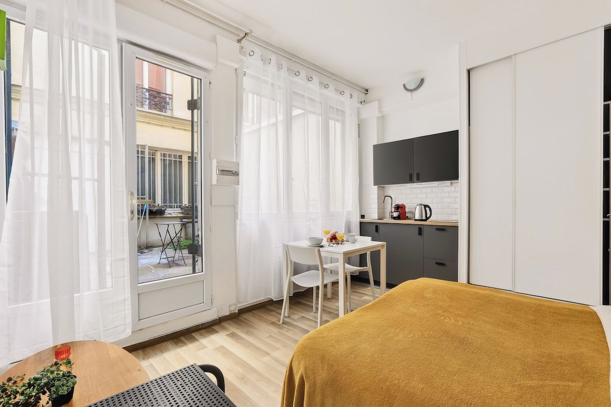 A compact room with a bed, a small table with a bowl and a cup, a kitchenette with a black cabinet, and a sliding glass door leading to an outdoor area.