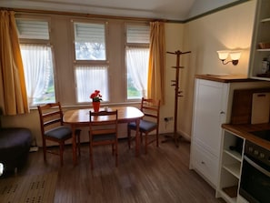 A dining area with a table and chairs, a window with curtains, a coat rack, and a kitchen area with cabinets and a stove. - Otto House - Holiday Apartment 1 (Wyk auf Foehr)