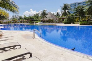 A resort pool with lounge chairs, palm trees, and a thatched-roof structure. - Beachfront Cancun Condo · Ocean View · Pool & Private Beach (Cancún)