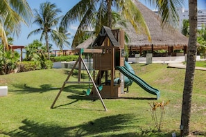 A wooden play structure with a slide, surrounded by grass and palm trees. - Beachfront Cancun Condo · Ocean View · Pool & Private Beach (Cancún)