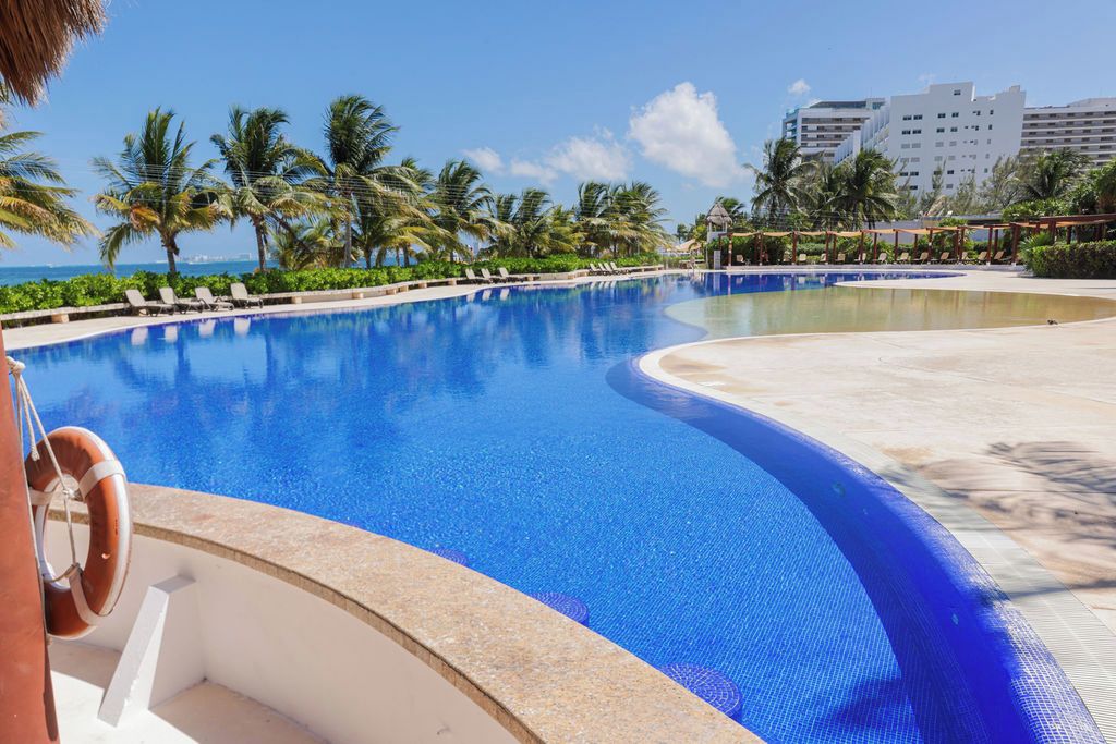 A large, clear swimming pool with a curved edge, surrounded by palm trees and lounge chairs, with a lifebuoy attached to the poolside.