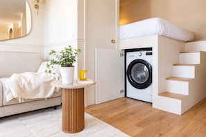 A compact living space with a sofa, a round table with a potted plant, a washing machine, and a built-in bed with a headboard. - New Listing (Paris)
