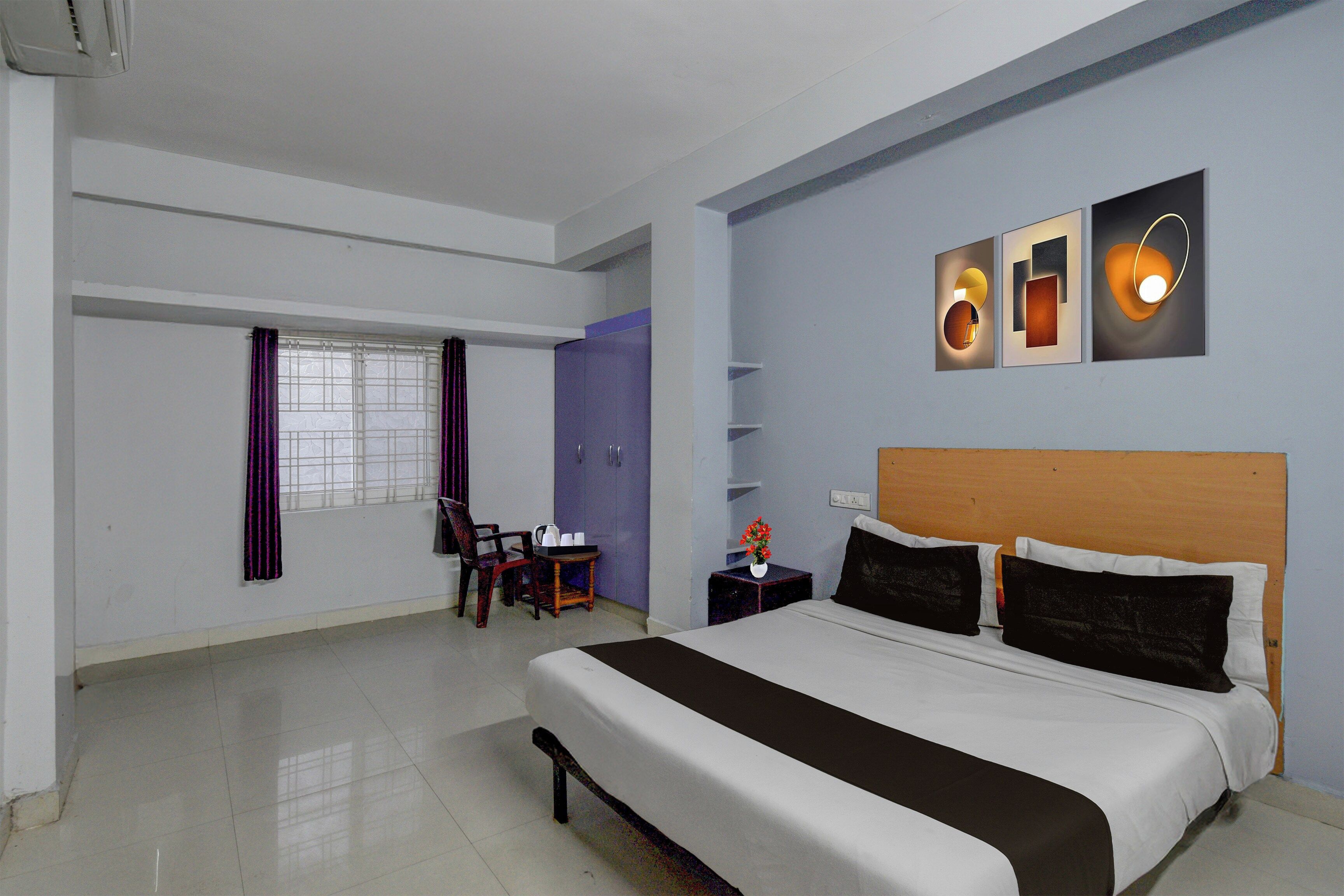 A hotel room with a large bed, a desk with a chair, a window with curtains, and wall-mounted artwork.