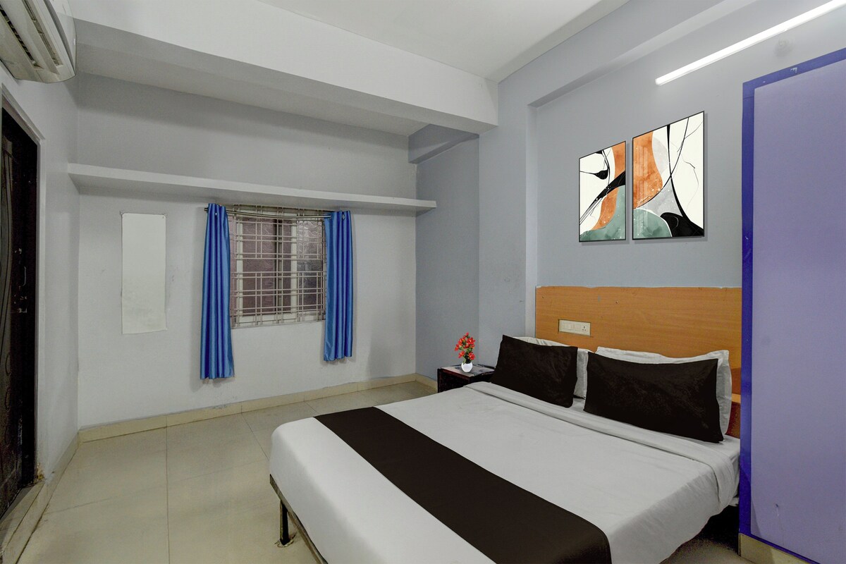 A hotel room with a bed, a window with blue curtains, a wall-mounted artwork, and a bedside table with a vase of flowers.