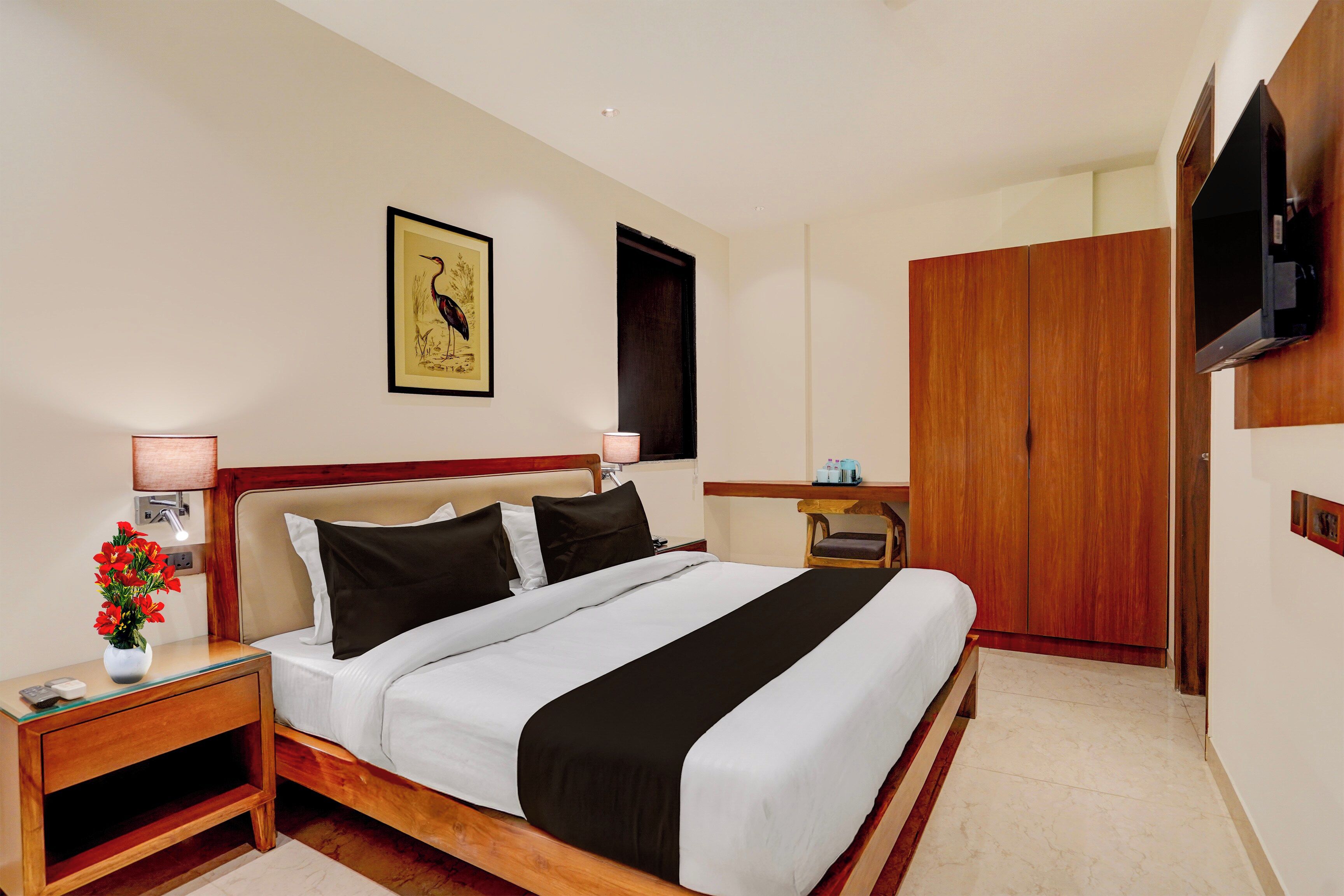 Habitación de hotel con cama, mesita de noche, lámpara, obras de arte, televisión, armario y una pequeña mesa con objetos.