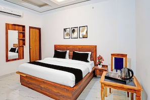 Habitación de hotel con una cama de madera, una mesita de noche con hervidor, un espejo y tres cuadros en la pared.