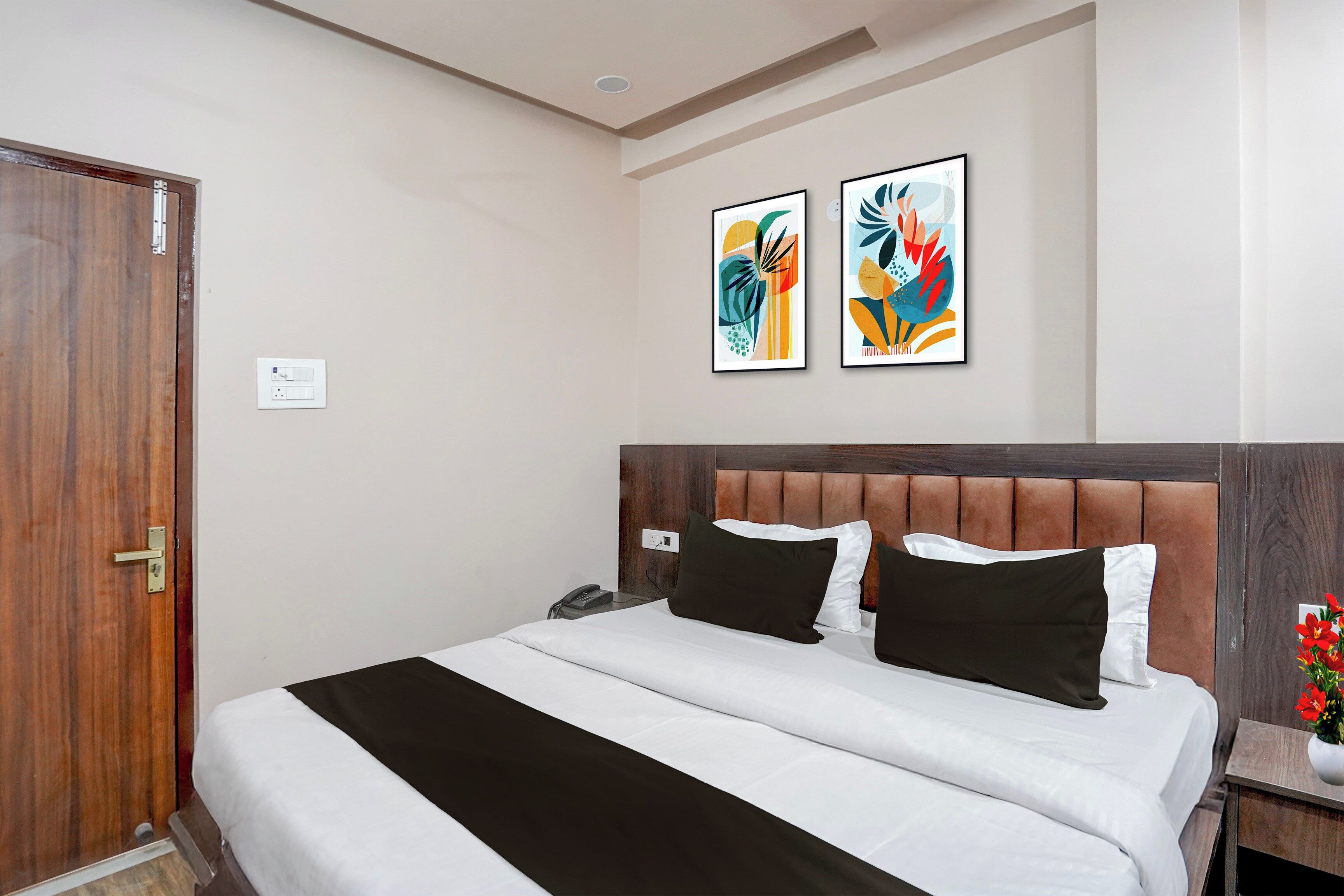 Una habitación de hotel con una cama, una puerta de madera, una mesita de noche, un teléfono y dos cuadros en la pared.